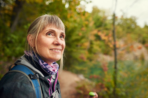 Senior woman wearing outdoor gear smiling and admiring the autumn scenery while out for a hike in a forest
