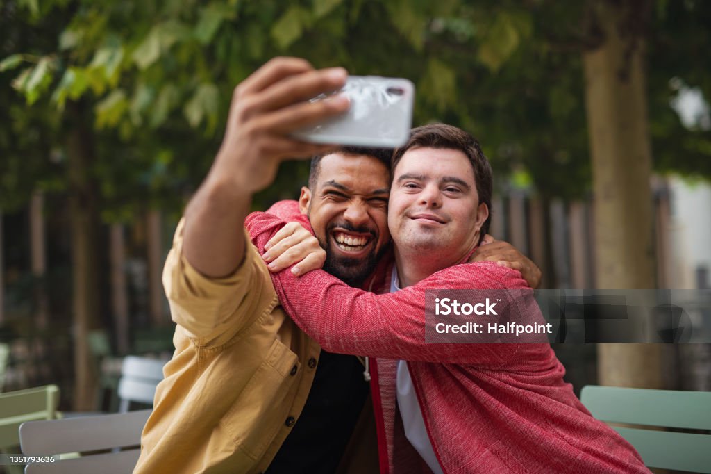 Young man with Down syndrome and his mentoring friend sitting and taking selfie outdoors in cafe A young man with Down syndrome and his mentoring friend sitting and taking selfie outdoors in cafe Down Syndrome Stock Photo