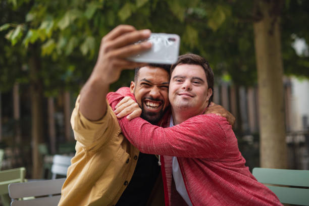 Young man with Down syndrome and his mentoring friend sitting and taking selfie outdoors in cafe A young man with Down syndrome and his mentoring friend sitting and taking selfie outdoors in cafe down syndrome stock pictures, royalty-free photos & images