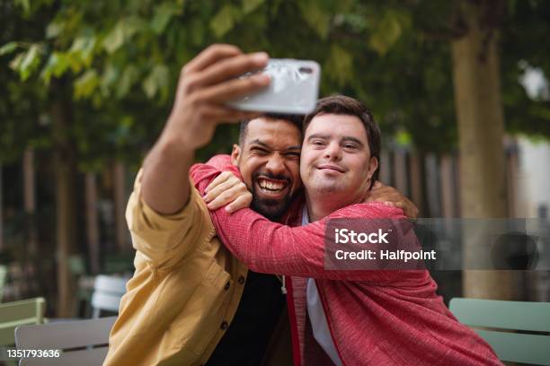 Young Man With Down Syndrome And His Mentoring Friend Sitting And Taking Selfie Outdoors In Cafe Stock Photo - Download Image Now