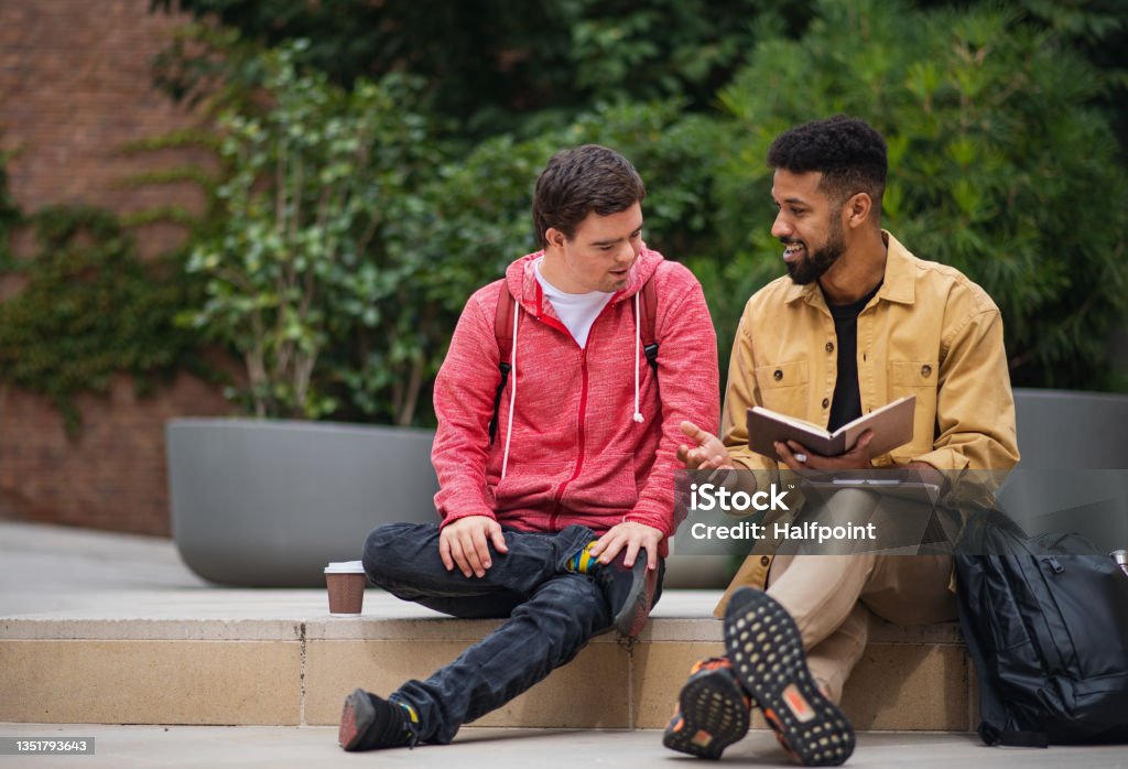 Happy young man with Down syndrome and mentoring friend sitting and talking outdoors A happy young man with Down syndrome and mentoring friend sitting and talking outdoors Disability Stock Photo