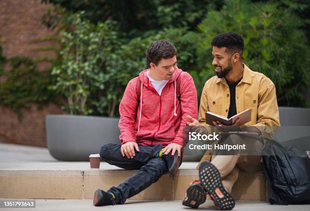 Happy Young Man With Down Syndrome And Mentoring Friend Sitting And Talking Outdoors Stock Photo - Download Image Now
