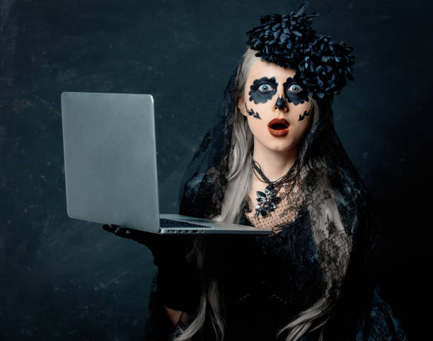 portrait of a girl in a witch costume with laptop computer on a dark background. Halloween holiday stock photo