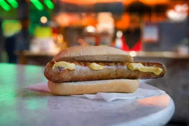 Bratwurst sausage with mustard in a bun, typical fast food at German traveling carnival, fair fun and Christmas market, blurred lights at night, copy space, selected focus, narrow depth of field