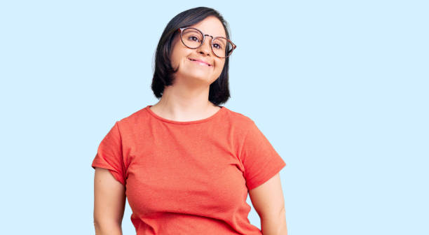 Brunette woman with down syndrome wearing casual clothes and glasses looking positive and happy standing and smiling with a confident smile showing teeth stock photo