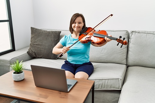 Brunette woman with down syndrome sitting on the sofa practicing violin at the living room