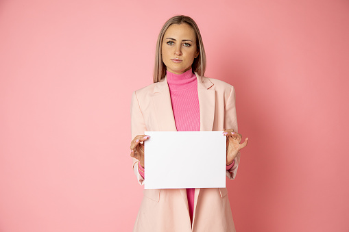 Portrait of attractive young blonde hair woman 30-35 years old holding blank of paper A4 for sign on pink background. businesswoman is wearing pink jacket. Top manager, female blog