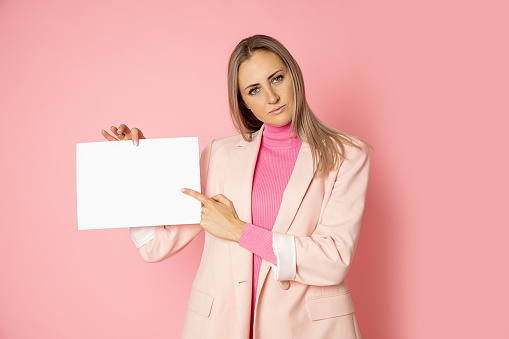 Portrait of attractive young woman holding blank of paper A4 for sign on pink background. businesswoman is wearing pink jacket. Points with finger at copy space.