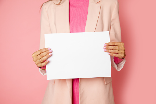 Close-up Photo of unrecognizable woman holding blank sheet of paper for commercial inscription. pink background. studio shot. for female blog article or website, social networks.
