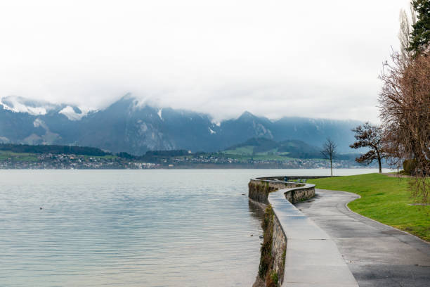 view of the lake Thun and the mountains wrapped in clouds a spring morning on lake thun, view from a small promenade with a view of mountain peaks behind the opposite shore of the lake. bernese oberland, Canton Bern, Switzerland lake thun stock pictures, royalty-free photos & images