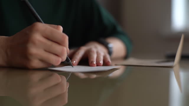 Close-up hands of unrecognizable young woman writing handwritten wishes on Christmas card sitting at table at home, selective focus.