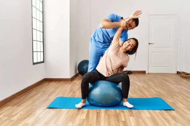 Latin man and woman wearing physiotherapist uniform having rehab session using fit ball at rehab center