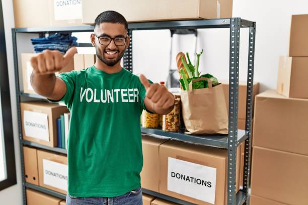 istock Young indian man volunteer holding donations box approving doing positive gesture with hand, thumbs up smiling and happy for success. winner gesture. 1351827154