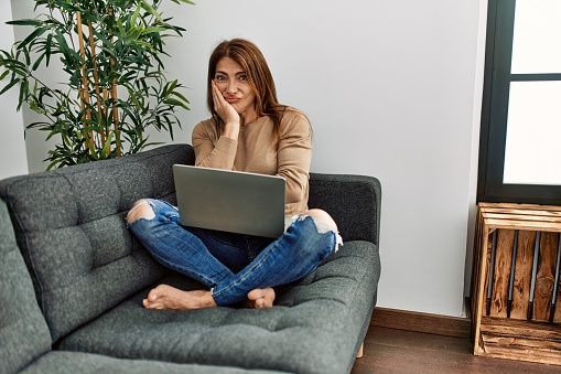 Senior middle east woman using laptop sitting on the sofa at home thinking looking tired and bored with depression problems with crossed arms.