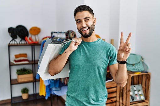 Young handsome man with beard holding shopping bags at retail shop smiling with happy face winking at the camera doing victory sign with fingers. number two.