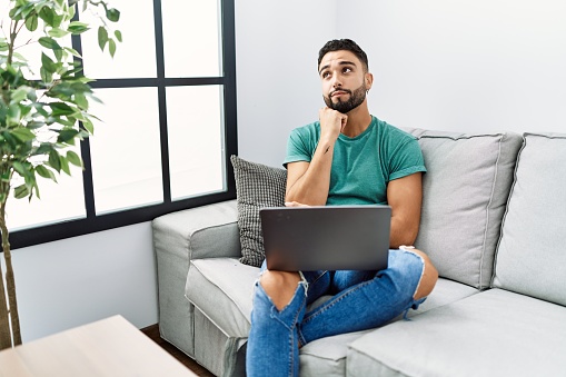 Young handsome man with beard using computer laptop sitting on the sofa at home with hand on chin thinking about question, pensive expression. smiling with thoughtful face. doubt concept.