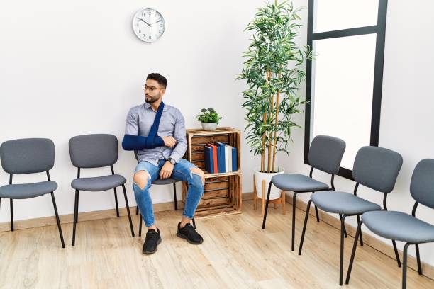 giovane uomo arabo che si siede sulla sedia con la fionda del braccio nella sala d'attesa della clinica - arm sling foto e immagini stock