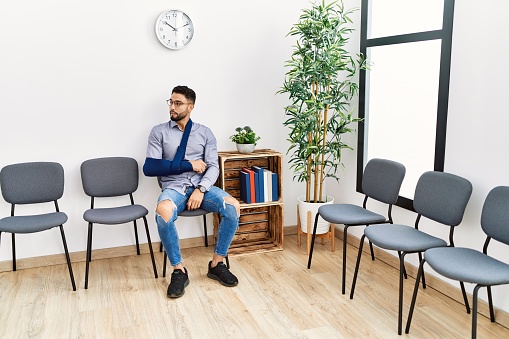 Young arab man sitting on chair with arm sling at clinic waiting room