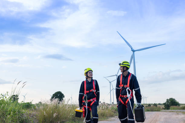 männliche und weibliche ingenieure in uniform, rote sicherheitsglocken, gelbe hüte, rutschfeste handschuhe für die arbeit in der höhe gehen in einen windkraftpark, um wartungsarbeiten durchzuführen. saubere energie aus der natur. - non rewnewable stock-fotos und bilder