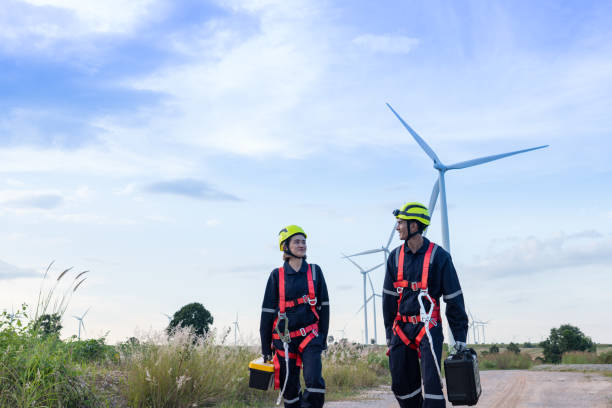 des ingénieurs hommes et femmes en uniforme, des cloches de sécurité rouges, des chapeaux jaunes, des gants antidérapants pour travailler en hauteur entrent dans un parc éolien pour effectuer l’entretien. énergie propre de la nature. - working windmill photos et images de collection