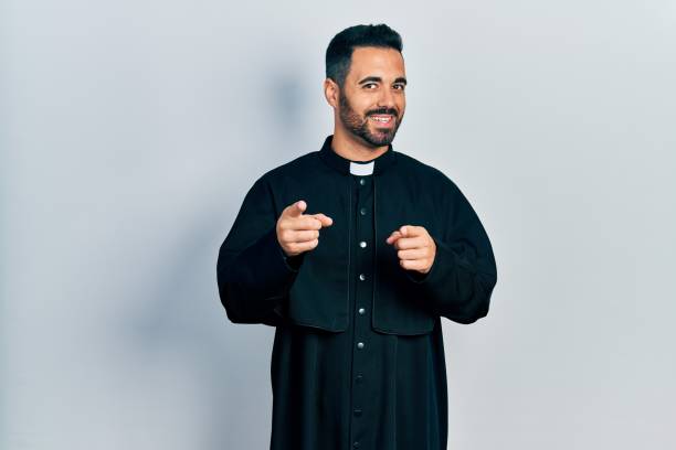 istock Handsome hispanic man with beard wearing catholic priest robe pointing fingers to camera with happy and funny face. good energy and vibes. 1351834976