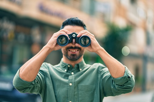 Young hispanic man smiling happy looking for new opportunity using binoculars at the city.