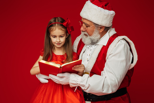 A cute little girl sitting on Santa Claus' lap and reading a book
