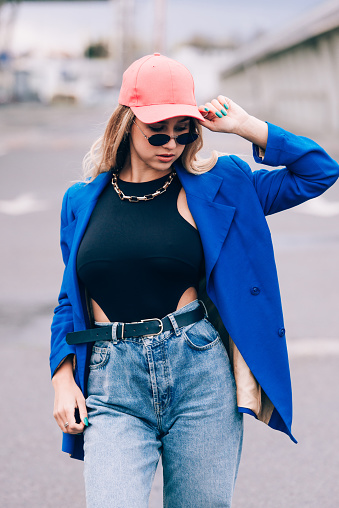 Young blonde hipster woman posing on the street. Wearing blue stylish jacket, jeans and baseball hat and sunglasses. Lifestyle portrait