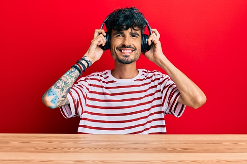 Young hispanic man using headphones sitting on the table smiling with a happy and cool smile on face. showing teeth.