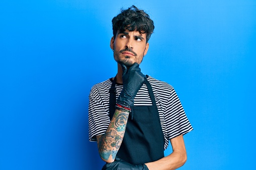 Young hispanic man wearing barber apron serious face thinking about question with hand on chin, thoughtful about confusing idea