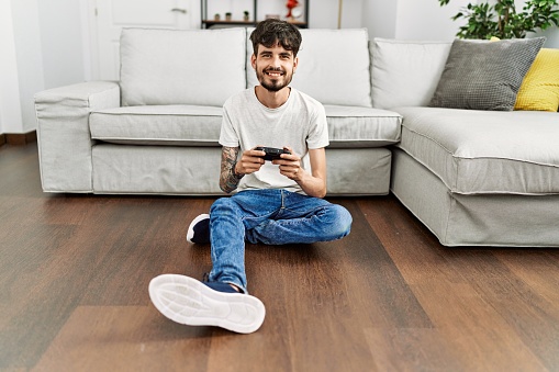 Young hispanic man playing video game sitting on the floor at home.