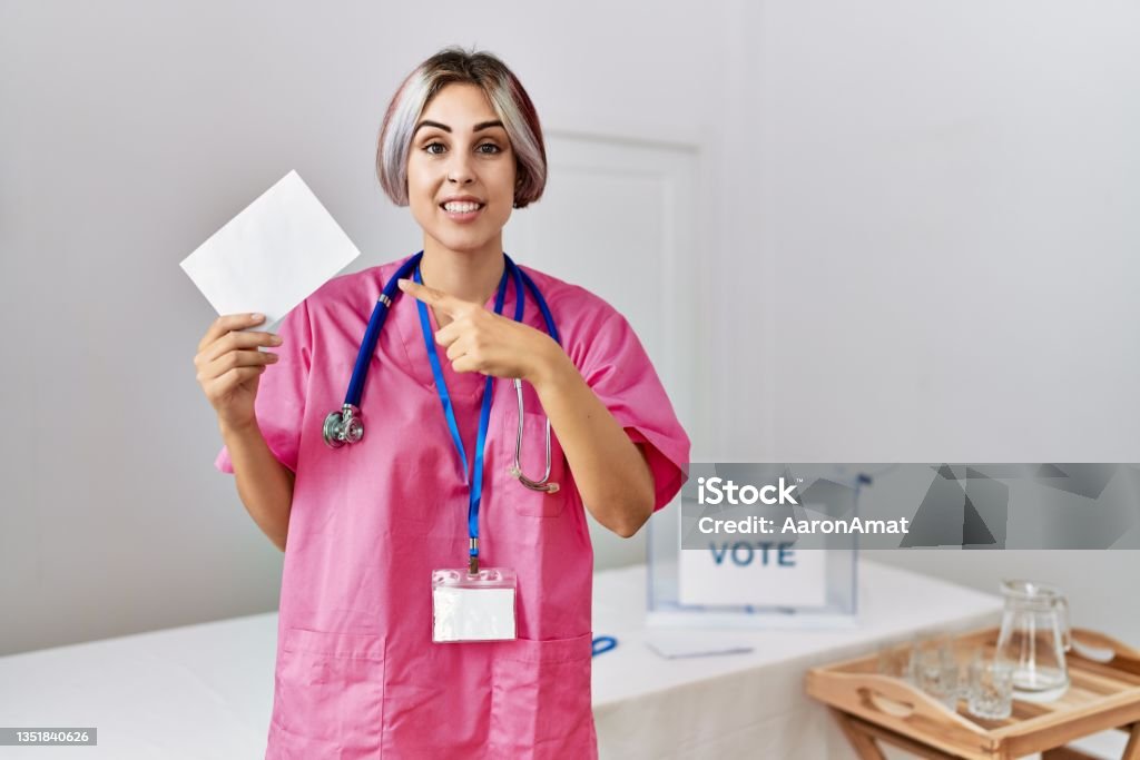Young nurse woman at political campaign election holding envelope smiling happy pointing with hand and finger Nurse Stock Photo