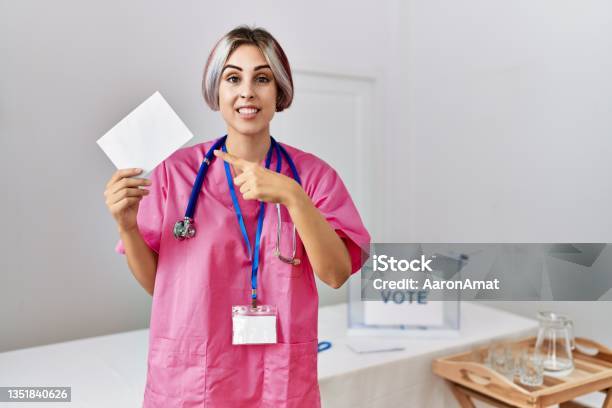 Young Nurse Woman At Political Campaign Election Holding Envelope Smiling Happy Pointing With Hand And Finger Stock Photo - Download Image Now