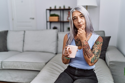 Middle age grey-haired woman drinking coffee sitting on the sofa at home looking confident at the camera smiling with crossed arms and hand raised on chin. thinking positive.