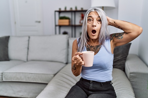Middle age grey-haired woman drinking coffee sitting on the sofa at home crazy and scared with hands on head, afraid and surprised of shock with open mouth