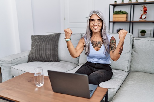Middle age grey-haired woman using laptop at home very happy and excited doing winner gesture with arms raised, smiling and screaming for success. celebration concept.