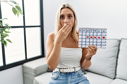 Young blonde woman holding heart calendar shocked covering mouth with hands for mistake. secret concept.