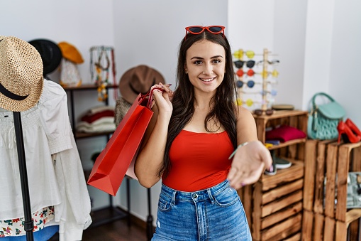Young brunette woman holding shopping bags at retail shop smiling cheerful offering palm hand giving assistance and acceptance.