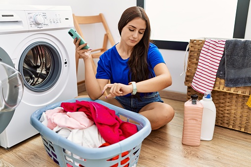 Young brunette woman doing laundry using smartphone checking the time on wrist watch, relaxed and confident