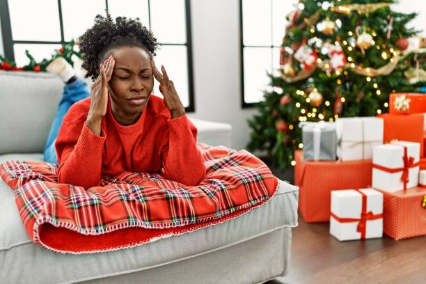 若いアフリカ系アメリカ人女性は、ストレスのために頭の痛みのために頭に手を置いてクリスマスツリーでソファに横たわっています。片頭痛に苦しんでいます。 - home  audio ストックフォトと画像