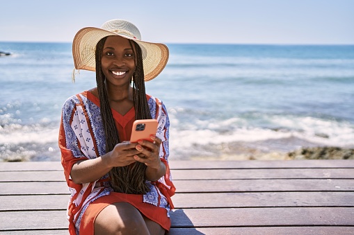 Young african american tourist woman using smartphone sitting on the bench at the beach.