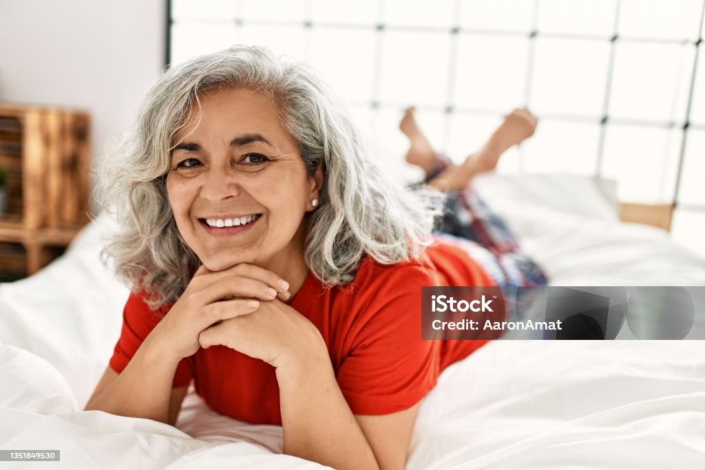 Middle age grey-haired woman smiling happy lying on the bed at bedroom. Mature Women Stock Photo