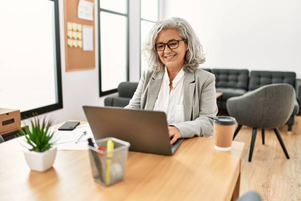 中年の灰色の髪のビジネスウーマンは、オフィスで働いて幸せそうに微笑んでいます。 - beautiful senior woman ストックフォトと画像