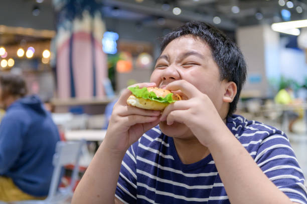 obese boy eats a hamburger in a food court in a shopping center. junk food - teen obesity imagens e fotografias de stock