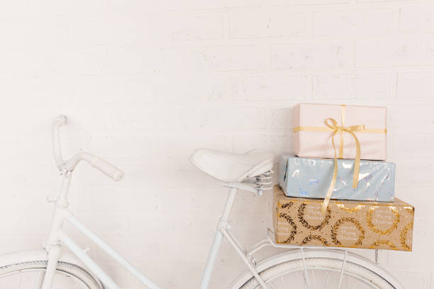 istock Original festive decor. White bicycle stands against background of white brick wall in back seat is pile of gift boxes. Wrapped in gift paper, tied with ribbons. Copy space. 1351850806