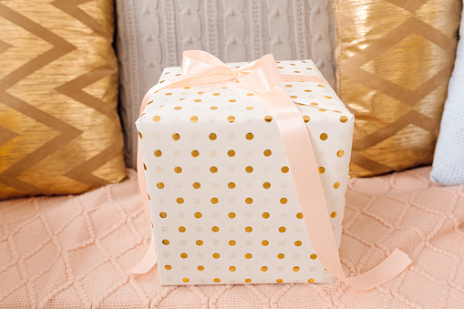 gift box wrapped in white gift paper with golden peas tied with pink ribbon on bow stands on pink background. Delicate pillows, surprise, gift concept, photo
