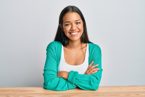 Beautiful hispanic woman wearing casual clothes sitting on the table happy face smiling with crossed arms looking at the camera. positive person.