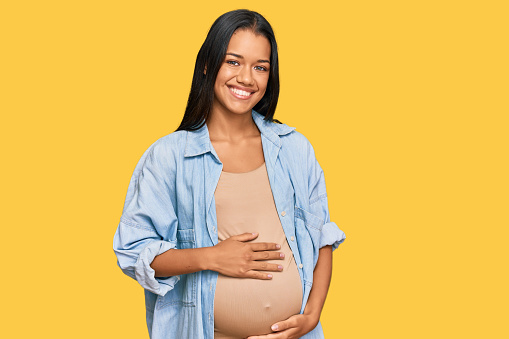 Beautiful hispanic woman expecting a baby showing pregnant belly looking positive and happy standing and smiling with a confident smile showing teeth