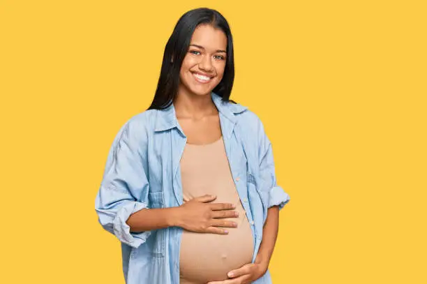Photo of Beautiful hispanic woman expecting a baby showing pregnant belly looking positive and happy standing and smiling with a confident smile showing teeth