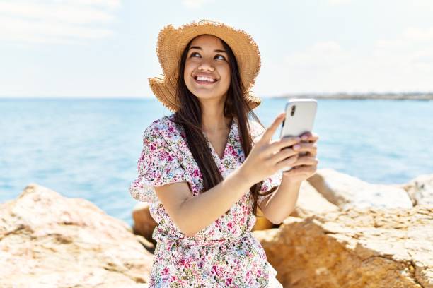 istock Young latin girl wearing summer hat using smartphone at the beach. 1351851383